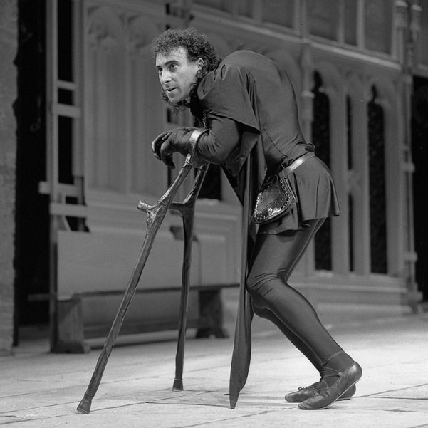 Against a backdrop of a windowed mediaeval wall, a very hump-backed Richard 3rd, clothed in a black tunic and black hose, leans on two crutches, his knees bent, staring intently to his (and our) left.