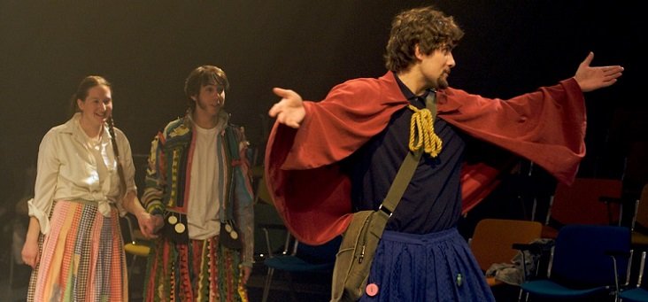 A man in rustic dress spreads his arms and is either appealing to someone or expressing despair. Behind him stand hand in hand a young man and a young woman in bright but not rich clothing.