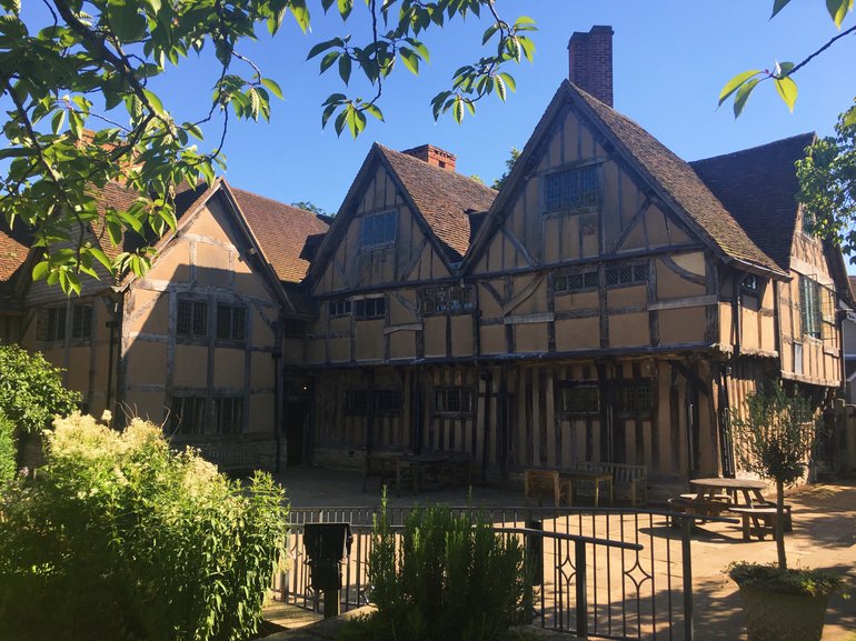 The back of Hall's Croft showing the patio and the three dormer gables forming two sides of the patio. The house is timber and brick, and has clearly just been restored. There is a gate from the patio to the garden, shrubs and a tree branch of which are at the left of the picture.