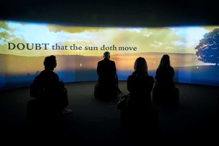 Image showing visitors in an exhibition space with low-level lighting, engaging with the immersive experience on a big screen that wraps around the room