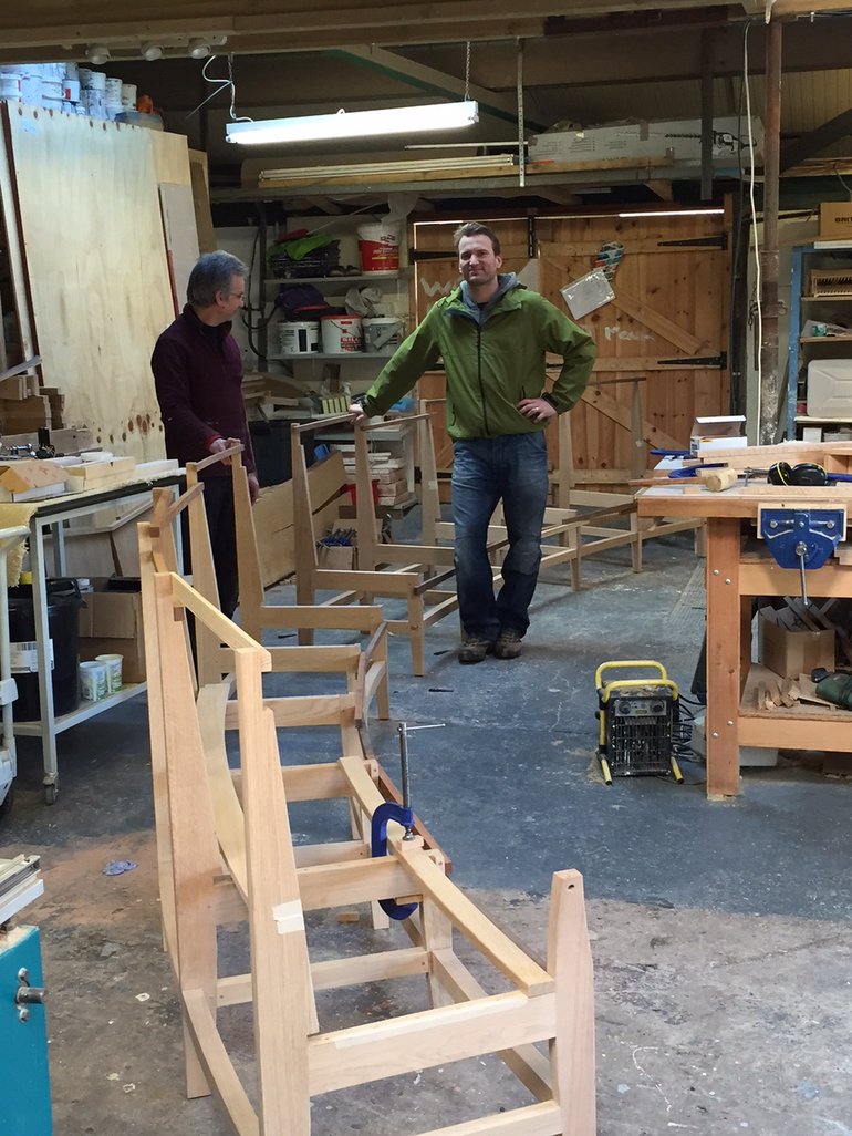 In the workshop, with tools and materials on shelves and workbenches, both men are looking at the curved run of seating benches, at this stage just frames with no seats.