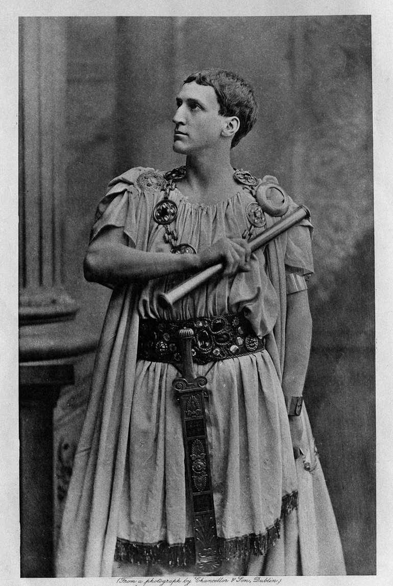 Sir Frank Benson as Coriolanus. He stands facing the viewer but looking upward to our left. He is wearing traditional Roman costume, with a wide belt from which a sword hangs centrally; he has a chain of office, and his right hand, holds a staff of office