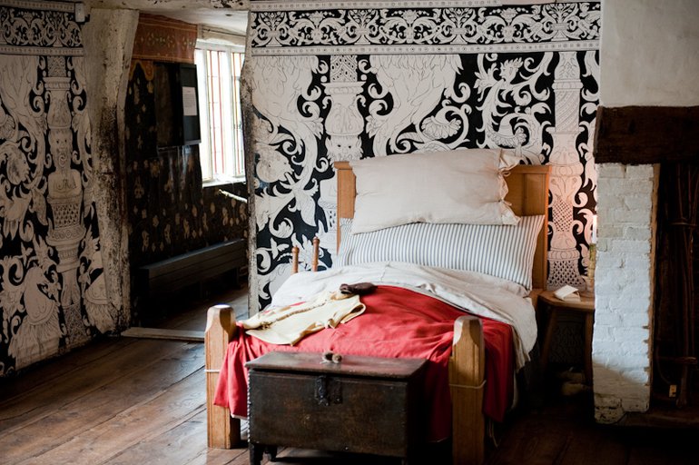 The cloths behind the bed and beside the window, are white with a black design of curving geometric shapes. The boys' bed has a red blanket, and a wooden chest stands at the foot.