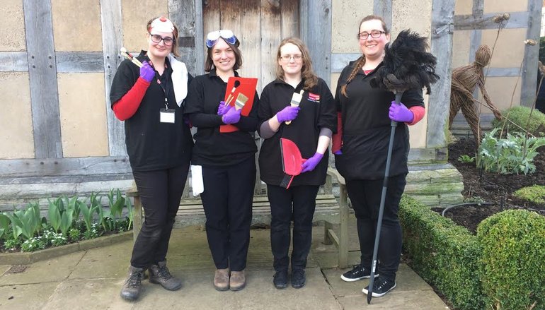 Standing in the garden with the Birthplace behind are four members of the conservation team in black overalls: a woman on the right has a feather duster, a woman on the left, small brushes, the two girls between them have other cleaning equipment.