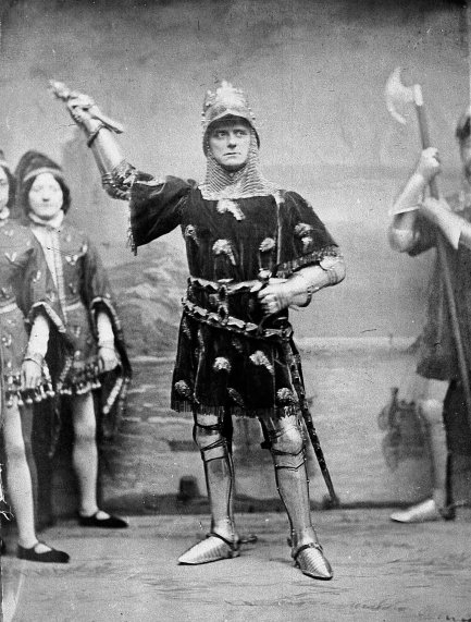 Charles Kean as Richard II, 1857. Helmeted and in armour with a black surcoat, he stands feet apart, grasping the hilt of his sword with his left hand, staring fiercely forward. His right hand is raised, holding what may be a dagger.