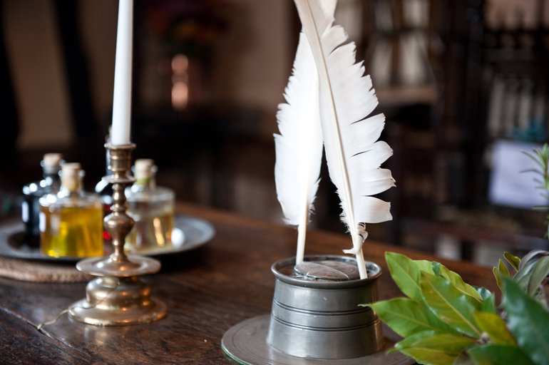 On a wooden table (against a blurred background) two white quill pens stand in a pewter ink pot. To the left is a white candle in a brass candlestick, behind which are three small corked bottles on a pewter (or silver) tray, one filled with dark liquid.