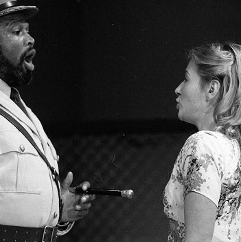 Emilia, blond-haired and wearing a short-sleeved dress, faces Othello, who wears military clothing.