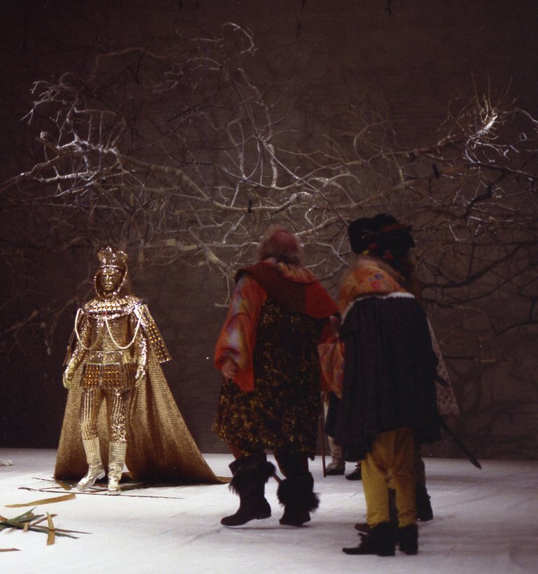 Two men with their backs to the camera, with red hair and wearing woollen tunics or cloaks, one with furred boots, stare at a figure in shining gold armour and a trailing cloak, with a crown on his head. Swords are on the floor, and a bare tree is behind.