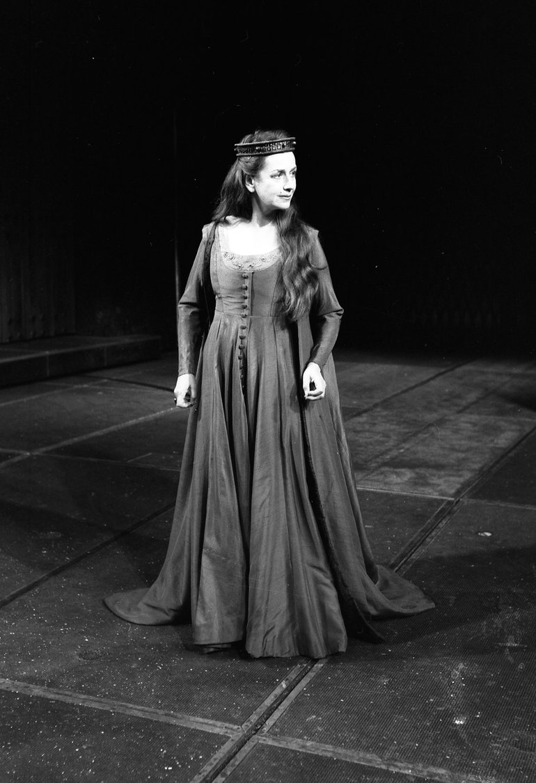 Queen Margaret stands facing us, but looking to her left. She wears a low-cut long dress with full skirt and long sleeves; she has long hair coming over her left shoulder; she wears a circlet, her hands are loose at her sides, and she has a half-smile.