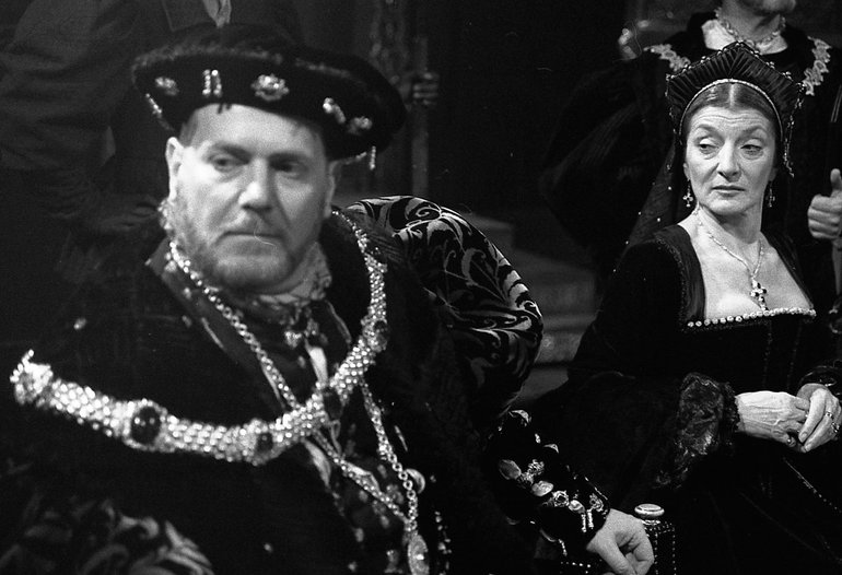 On the right, Queen Katherine, wearing black and an elaborate headdress looks disparagingly at Henry on her right. He, wearing many jewels, leans away from her; he looks stern, but a little troubled.