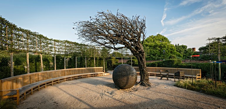 Within a semi-circle of benches topped on the left with espalier plants  on poles, A bare tree curves over a large stone ball.