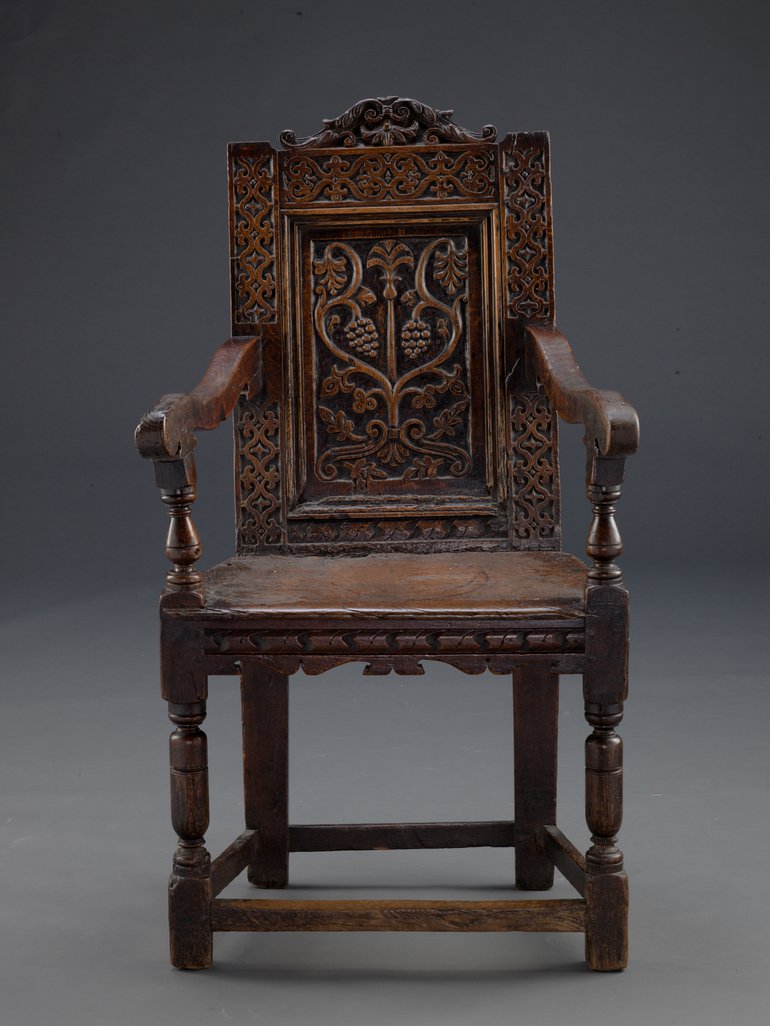 A portrait image of the Falcon Inn chair. The chair is made out of dark wood, the back of the chair is finely carved. It looks similar to the kind of chair you might still find in an old inn or pub.