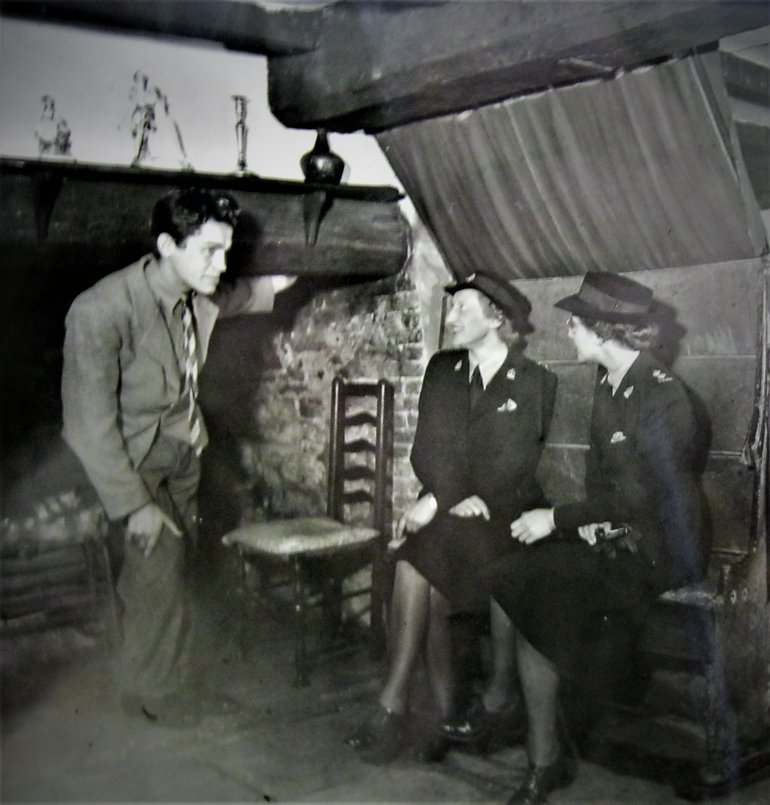 A black and white photo of two Canadian Nurses sit on the Courting Settle. They appear to be engaged in conversation with a man dressed in suit. He is standing in front of a fireplace.