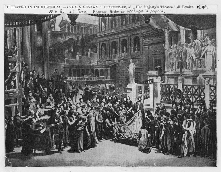 Against an impressive backdrop of tall classical buildings, standing on the steps of a plinth adorned with classical statues, Mark Antony speaks to a large crowd.