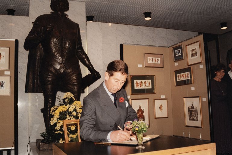 King Charles III, then Prince Charles, signing the visitor book in the Shakespeare Birthplace Trust's Marble Hall. He is sat at a table in front of a statue of William Shakespeare.