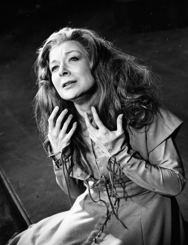 King John, RSC, 1957. Constance sits with her body twisted, her hands with spread fingers at her throat, and her loose dark hair cascading over her shoulders. She is wearing a 1940s raincoat.