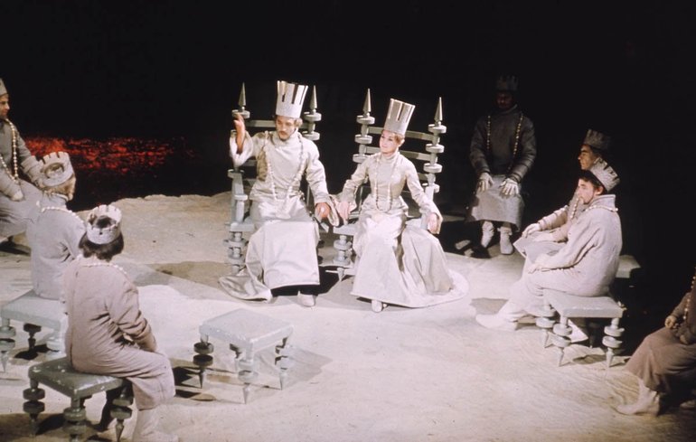 Macbeth Royal Shakespeare Company, 1967 (featuring funny hats). All characters are dressed in white. Macbeth and his lady sit on high-backed chairs facing us, other characters sit on stools around and facing them. All wear paper or cardboard crowns, Macbeth's and his lady's being taller than the rest.