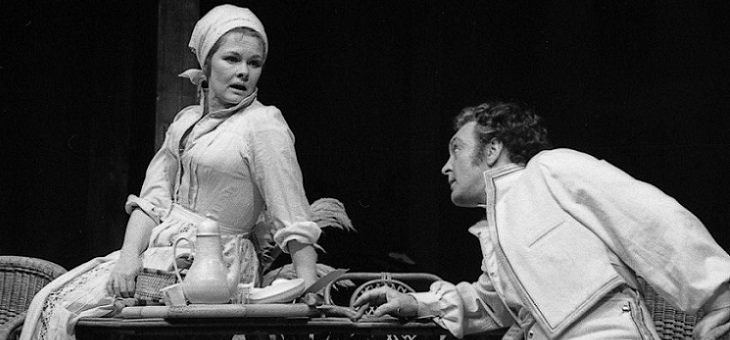 Beatrice, sitting on a garden table and resting her left arm on it, looks down at Benedick, who is leaning with his right forearm on the table and looking slightly hopefully up at Beatrice.