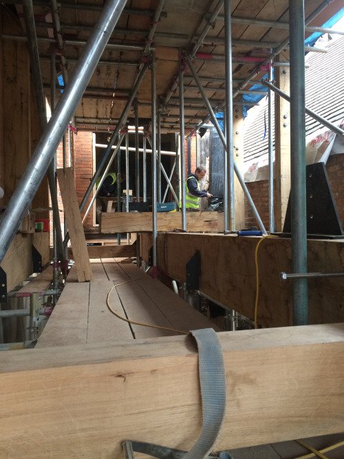 Nash's House Conservation. A two-level scaffolding and board construction from which towards the far end a man is seen doing something to the wall of the house.