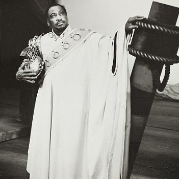 Othello stands with his left hand resting on part of a ship. He cradles a helmet in his right arm.