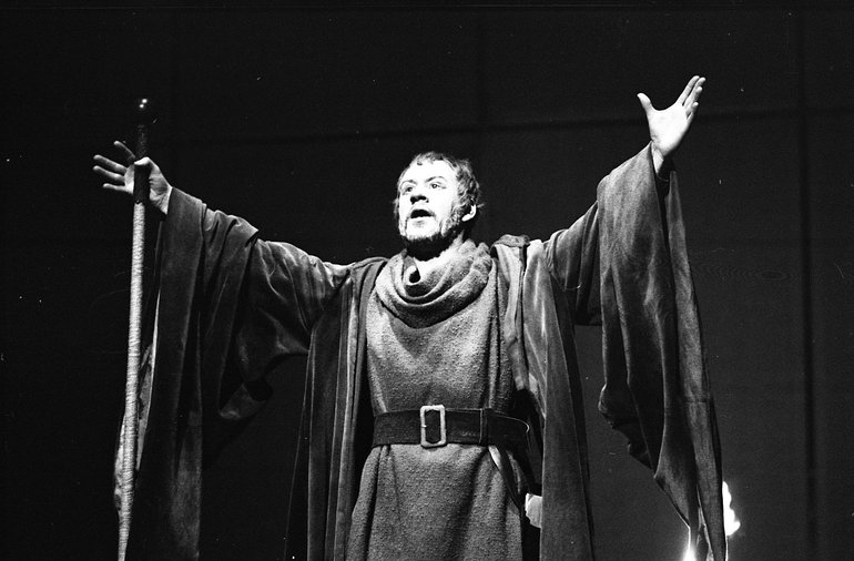 Pericles, Prince of Tyre, RSC, 1969. Spotlit against a dark background the character holds his arms wide and looks upwards, appearing to be praying or welcoming someone unseen. He wears a long woollen robe, a gown with long hanging sleeves, and he carries a stick.