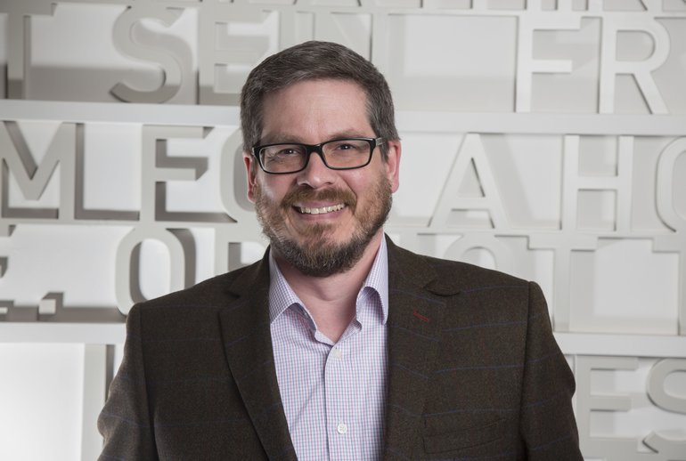 A photo of Nic Fulcher - Young(ish), dark hair and short beard, spectacles, smiling, wearing a dark jacket and an open-necked shirt.