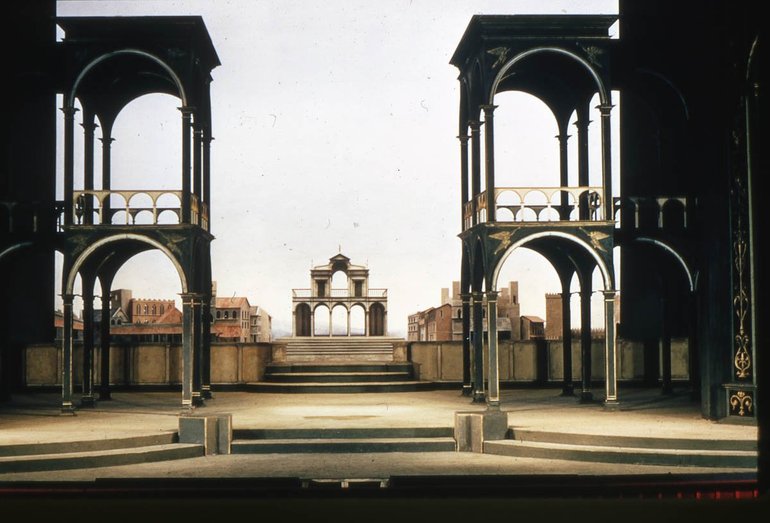 The empty set: a platform two steps above the front stage has matching structures each side. Each is an arched arcade with pillars, with the same above but with an open arched low balcony rail. To the rear are some steps, and a higher platform with some low buildings and a distant arched structure in the centre.