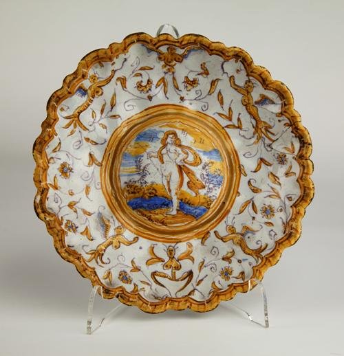 The plate has at its centre a naked figure in a landscape; concentric brown and gold rings surround this, outside which a brown design on the white ceramic has strange half-human figures, plants and marine-appearing motifs . The deckled edge is brown.