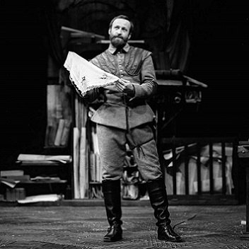 Iago is bearded, and wears a military uniform with knee-length boots. He is holding a strawberry handkerchief.