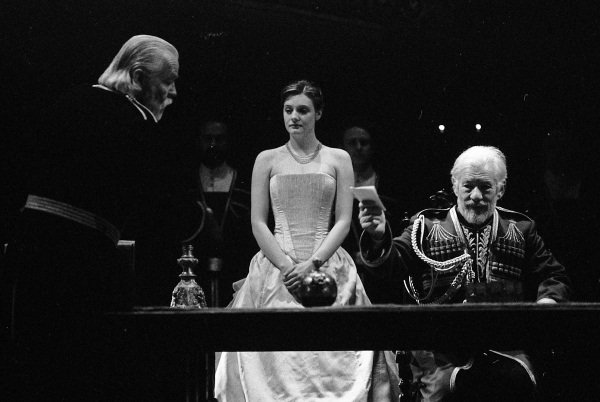 Cordelia in a white dress, her arms bare stands uncompromisingly staring at the white-haired Lear. A man in military uniform kneels beside Cordelia.