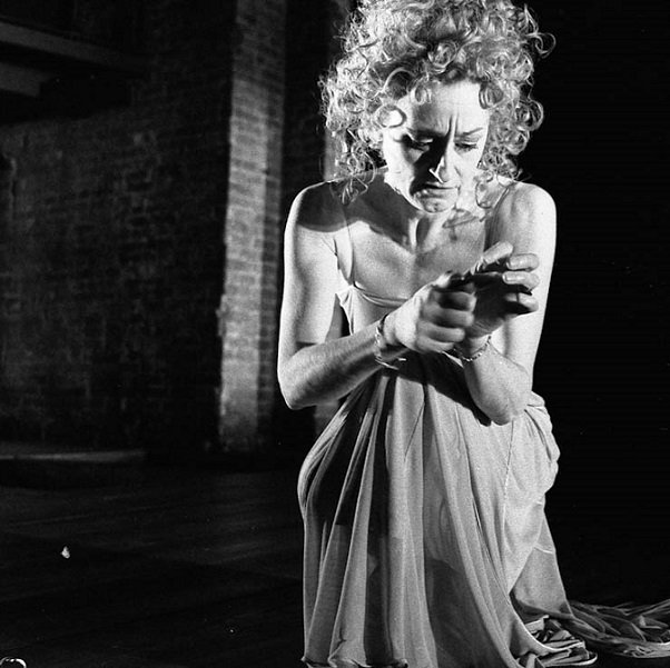 Spotlit against a dark background, a tortured Lady Macbeth with a mass of disordered curly hair hanging to her shoulders, and clad in her nightclothes, is washing her hands. (2004 production)