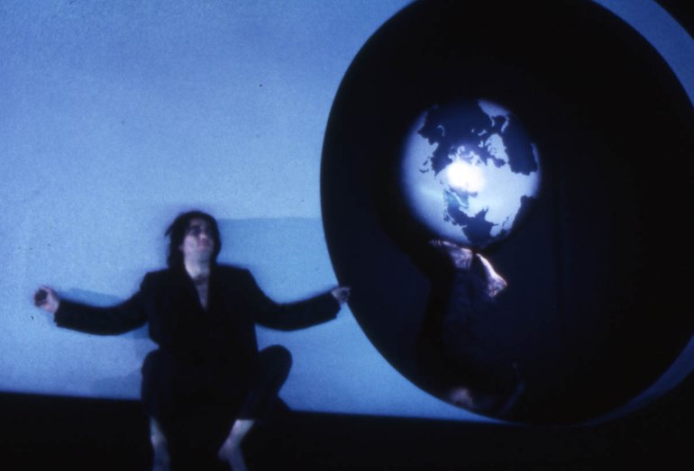 A man sits against a white wall with his arms spread out. Next to him is someone, wearing black clothes against a black background, balancing a globe on the bottom of their feet.