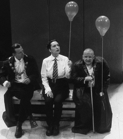 Simon Russell Beale sits on a bench wearing a black gown, leaning on a walking stick and holding a balloon. Beside him sit two younger men, one in shirtsleeves with a tie, and holding a balloon as well, the other in an open jacket.