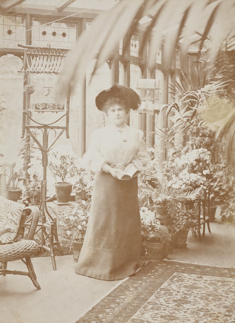 A black and white photograph of a lady in a hat, inside a conservatory filled with plants.