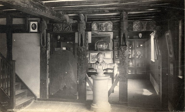 A bust of Shakespeare stands on a pedestal. A doorway on the right leads to a room with showcases of relics, which can also be seen through missing wall-panels behind the bust. A staircase is on the left.