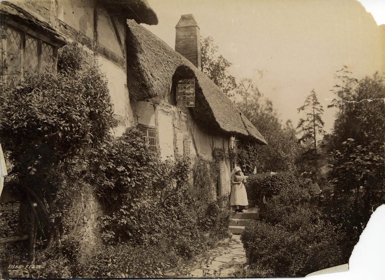 The picture show the oath and steps leading to the front door. To the right of the path are some tall, dark shrubs, while on the left bushes grow up the wall of the cottage. Mary Baker stands outside the front door, quite a distance; she wears a white head covering, a grey dress to her shins with a white apron over it, and she appears to be holding an open book in her hands.
