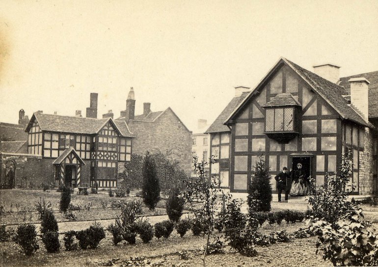 A black and white photo of the gardens at Shakespeare's Birthplace in 1870s. A man in a Derby hat is helping a lady in a period dress down the step into the garden.