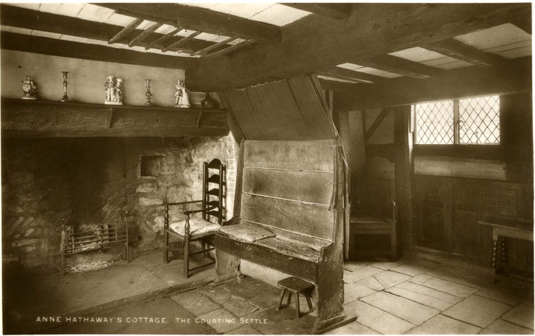 The very plain settle has a tall back, the top section of which slopes over the seat. It stands near the fireplace in the kitchen. On the hearthstone beyond it is an armchair and there is a small four-legged stool under the settle. The kitchen has a stone floor, and a lattice window on the right.