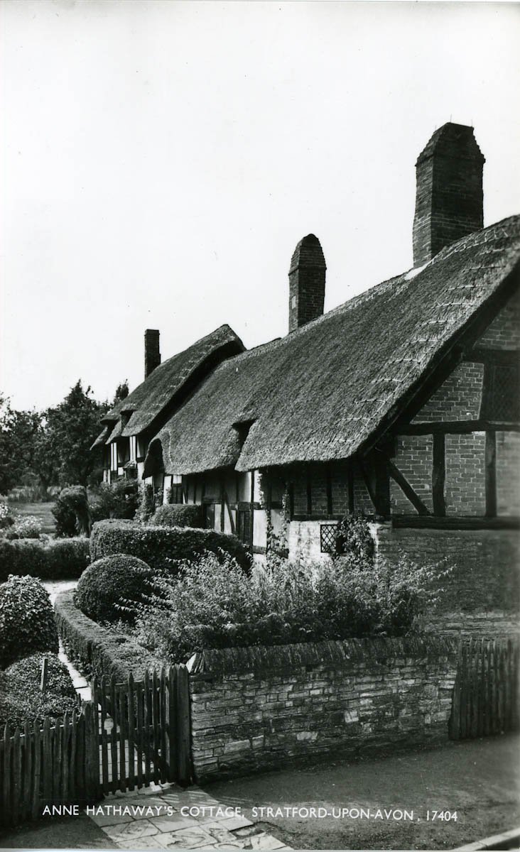 A postcard of the cottage, black and white, taken from the road and showing the garden gate and a path between neatly-trimmed hedges leading to the front door. The cottage itself looks very smart; the front is very shaded by the overhang of the thatch.