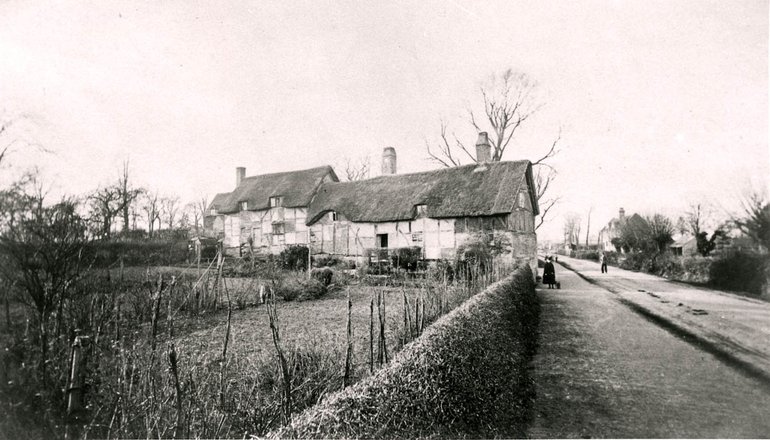 A low wall divides the garden from the road, with a random series of stakes behind it. A lawn is divided from the next lawn by a ragged hedge leading towards the cottage in the background..