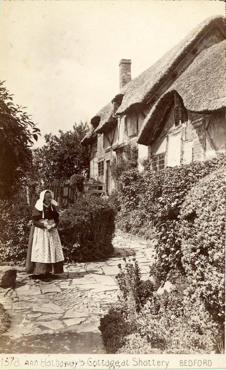 Mary Baker stands on the path in front of the cottage. She wears a white apron over a long dark dress, and her white head-covering which rests on her shoulders looks much like a nun's wimple. the path is edged on both sides by tall shrubs.