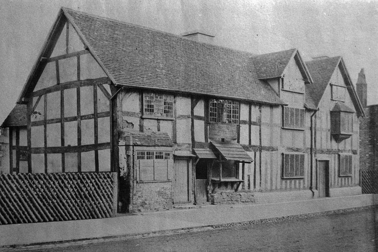An 1858 photo of the Birthplace. It shows doors to each side of the house, a shop counter and shop door in the first house with a fixed canopy over each, and the further house has a first-floor bay window. That house, with the gable ends facing the street, appears to have brick replacing wattle nd daub, and to be in much better condition than the nearer ppart with the shop.
