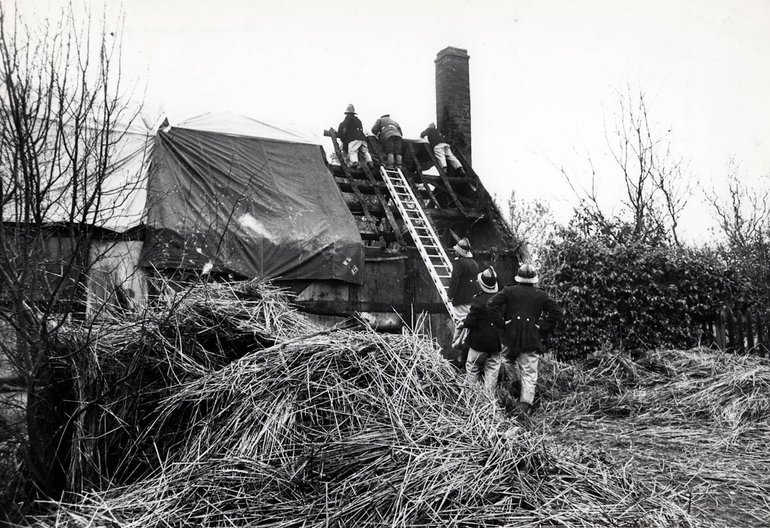A ladder runs to the top of the road end of the cottage, and at the top three firemen are placing a tarpaulin over the far side of the roof. To their left, both sides of the roof are covered with tarpaulins. At the foot of the ladder are three more firemen, and the ground around them is covered with piles of straw.