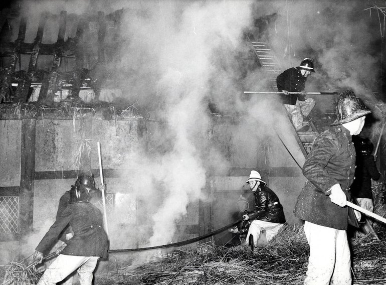 Behind a large plume of smoke and steam, the cottage is just visible, notable the roof timbers with no thatch. In the foreground two firemen with a hose are trying to douse the thatch; another, actually standing on the house wall, is raking the thatch down. A fourth fireman at the extreme right is raking something on the ground.