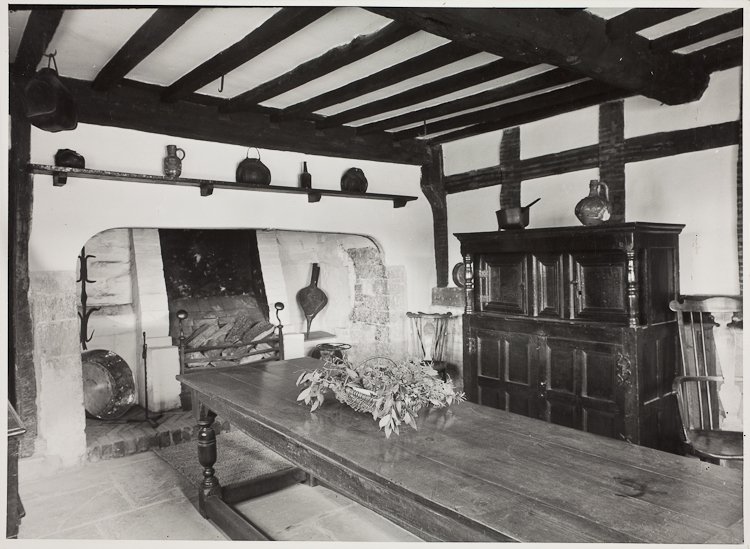 In the foreground is a long wooden table, at the back the large fireplace with a smaller grate set within it. There is a wooden dresser on the right, with an open shelf above cupboards and the top supported at the front by pillars.