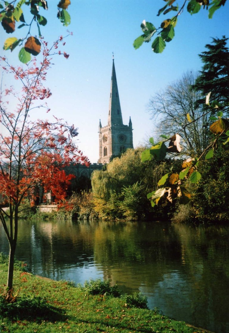 Across the river, the Tower and spire of Holy Trinity church. The bulk of the church is hidden by trees along the far river bank, and a sapling with red foliage is in the foreground on the left.