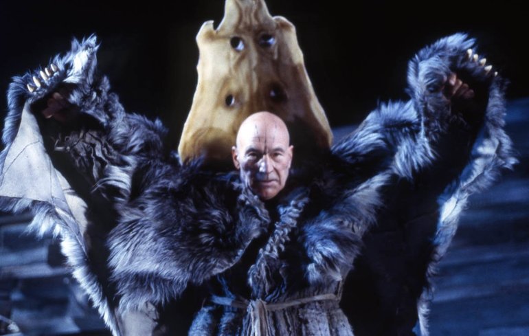 The Tempest Royal Shakespeare Company, 2006. Standing in front of a rock, a grim-faced Prospero holds his hands wide above his head. He is dressed in a black robe, over which he wears a furred cloak; held in his hands, this gives the impression of wings.