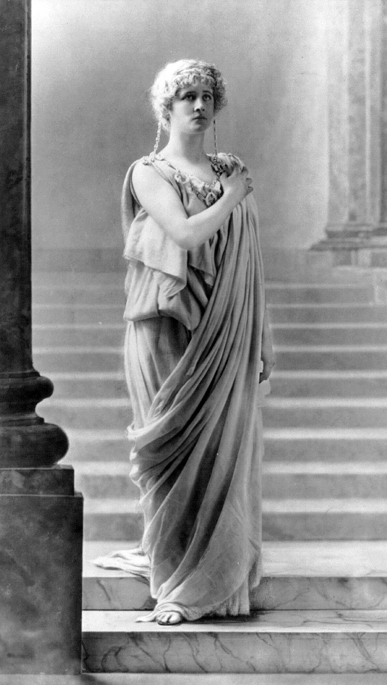 Dressed in a flowing robe, she stands on steps framed by pillars, her right hand on her left shoulder. She looks indeed like a statue.
