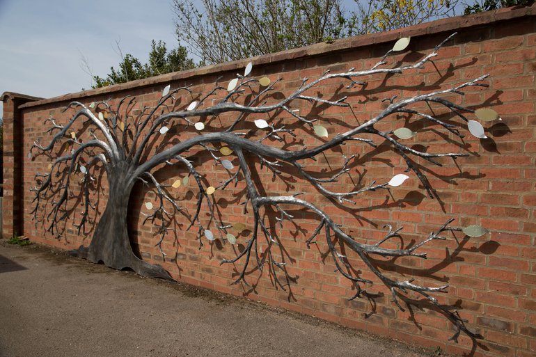 The tree is steel-coloured, its trunk flattened, and it spreads its branches in both directions along  brick wall, but with the branches to the right significantly longer.