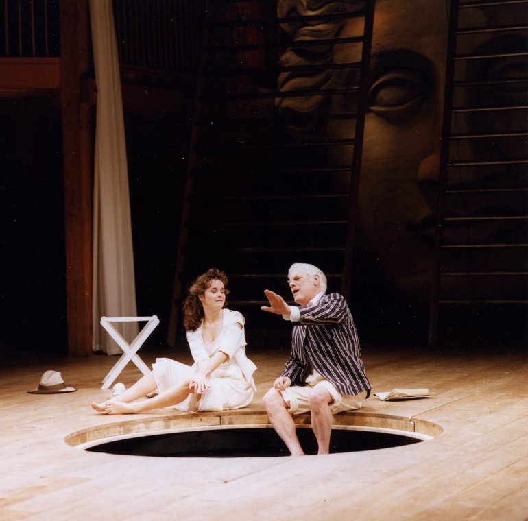 In a modern white dress, Cressida sits bare-legged by a pool. An older man, wearing a striped blazer and white shorts, sits with his bare legs in the pool. He is explaining something to Cressida, and his left hand is stretched out in front of him.